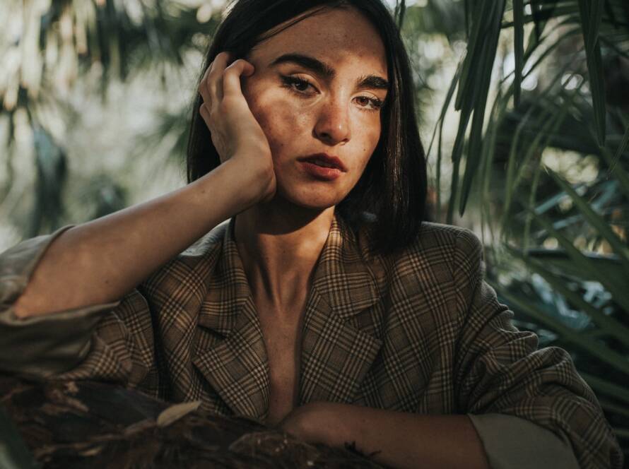 A portrait of a woman in a plaid blazer leaning on her hand, plants are in the background.