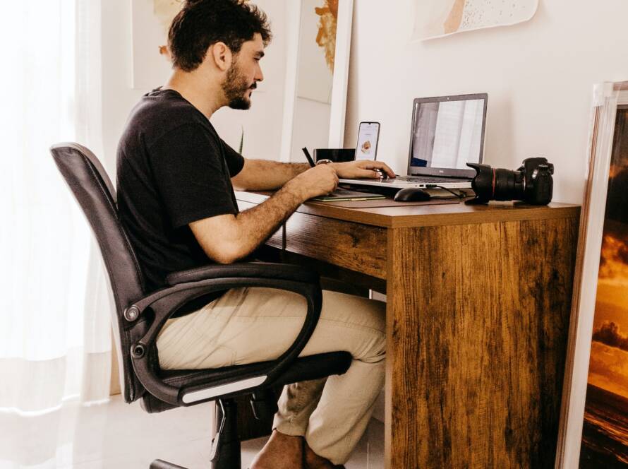 A man works on his laptop at a desk with a camera and phone nearby in a home office.