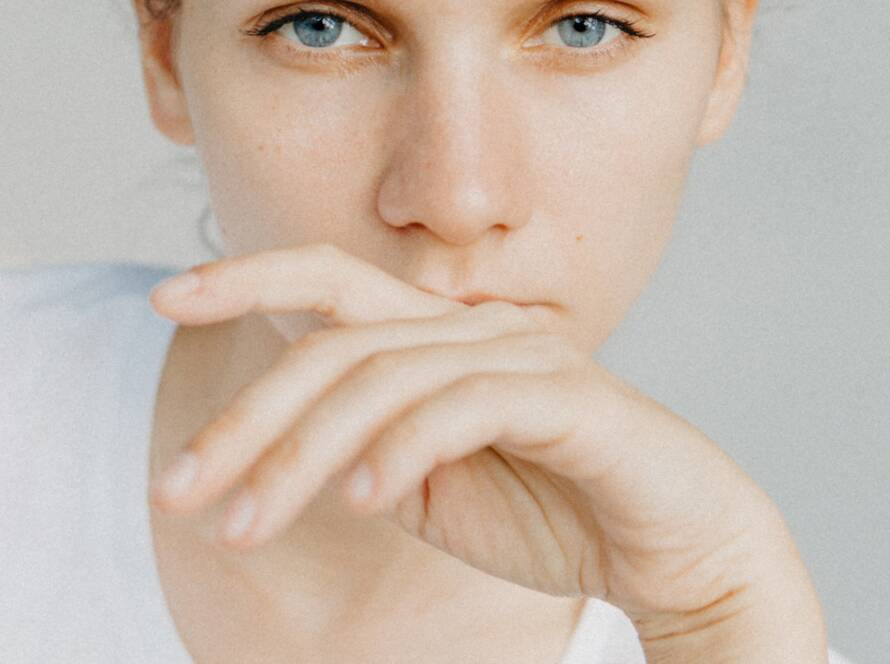 A thoughtful woman with blue eyes holds her hand near her mouth, looking directly at the viewer.
