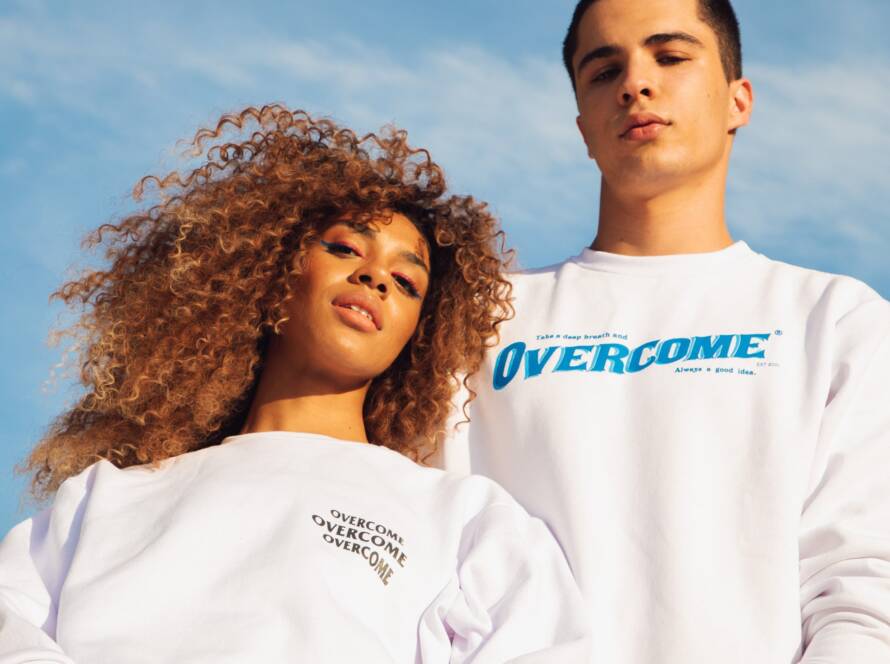 Two people wearing white sweatshirts with the word "OVERCOME" on them, standing in front of a blue sky.