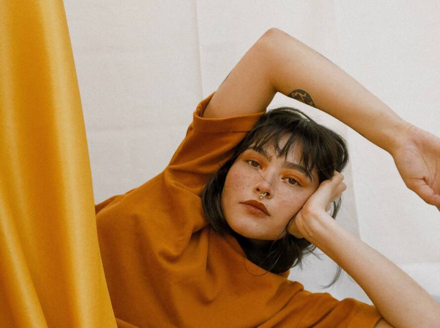 A woman wearing an orange shirt, with freckles and a nose ring, poses with one arm up and a hand on her face.