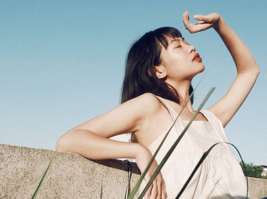 Young woman with closed eyes, hand up, shielding from the sun. She is outside and wearing a white top.