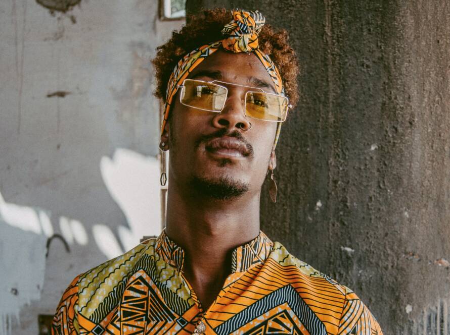 A Black man with glasses and a headband poses against a textured wall, wearing an orange and yellow patterned shirt.