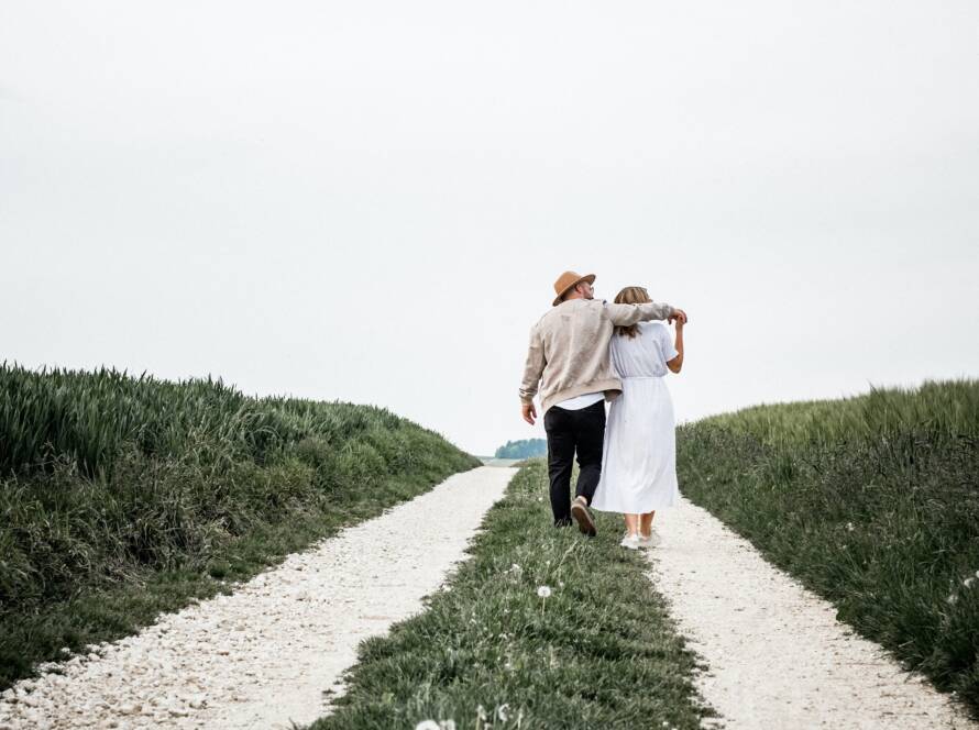 Couple walking away on a gravel path with arms around each other, surrounded by fields under a cloudy sky.