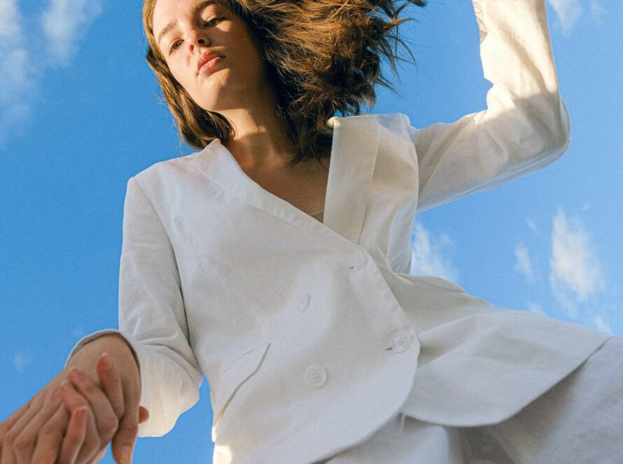 A young woman in a white suit looks upwards against a bright blue sky with wispy clouds.