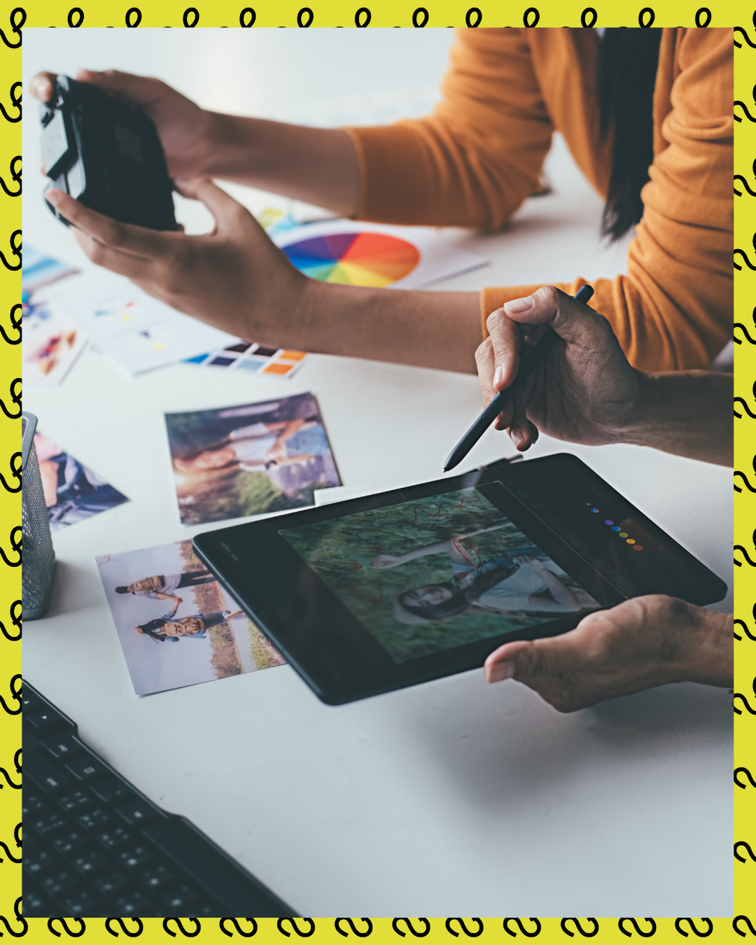 Two people working on a tablet editing photos, with a camera and printed photos on the table.
